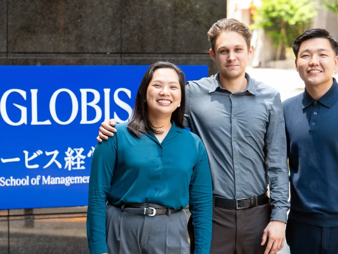 Three diverse, smiling students posing together in front of a blue GLOBIS University sign