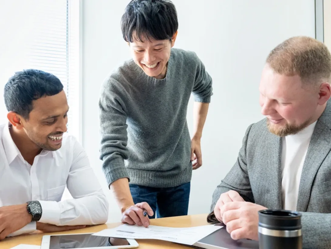 Three smiling male students stand around a table with documents in a classroom