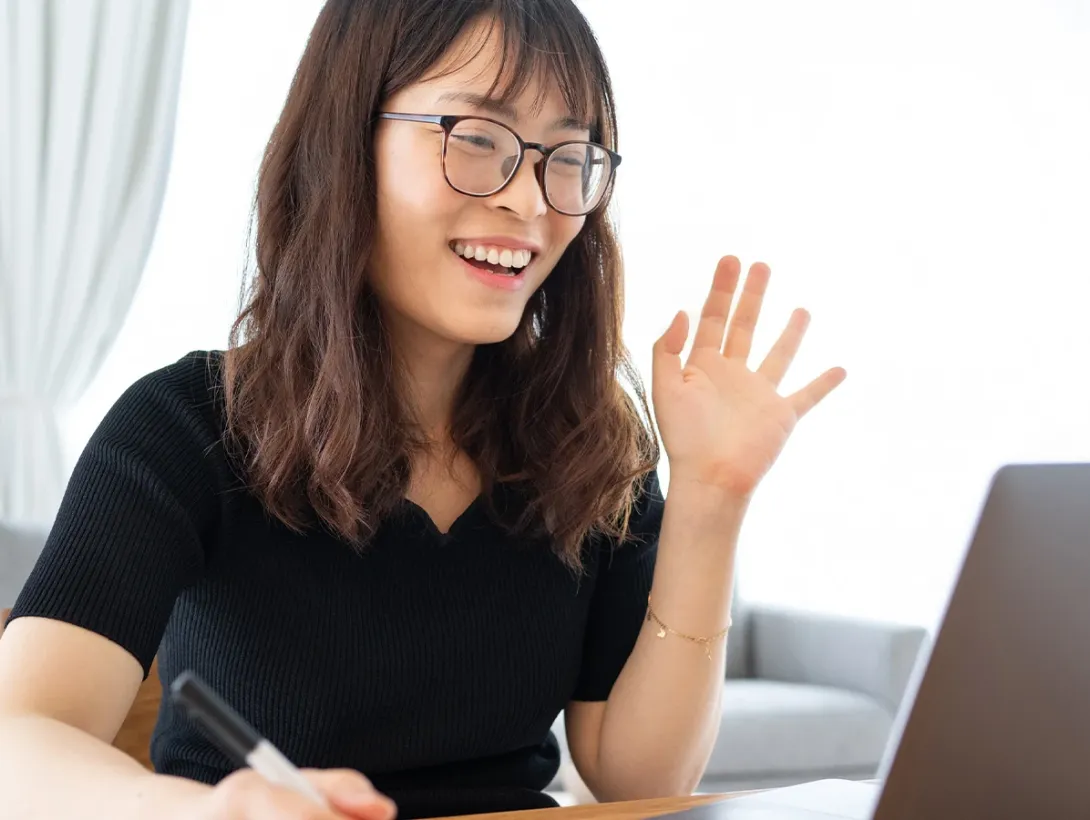 A young woman wearing glasses smiles and waves into a laptop camera while taking notes during a virtual class