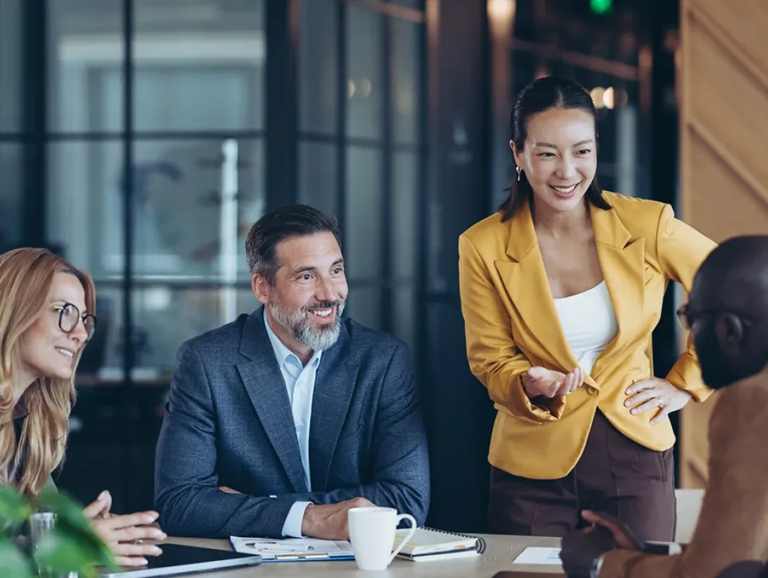 A woman standing leads a diverse group of smiling professionals in a collaborative discussion.