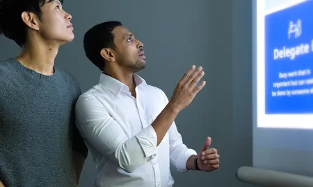 Two MBA students stand in a modern learning space, focusing a digital presentation projected onto a wall. One student gestures toward a slide, explaining a management framework to his peer.