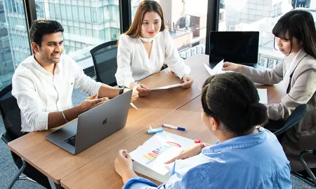 Four diverse students sit around a table in a high-rise office, discussing an assignment during a breakout session.