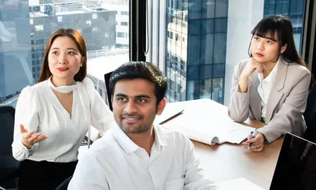 Three young professionals sitting at a conference table in a high-rise classroom, engaged in a collaborative group discussion.