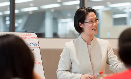 A women holding a marker and leading a presentation next to a whiteboard with business notes in a modern campus space.