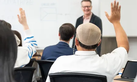 Students in a business lecture raising their hands to answer a question from the instructor standing by a whiteboard.