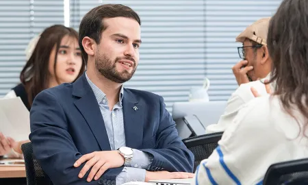 A man in a blue blazer actively participating in a group discussion with classmates in a bright, modern classroom.