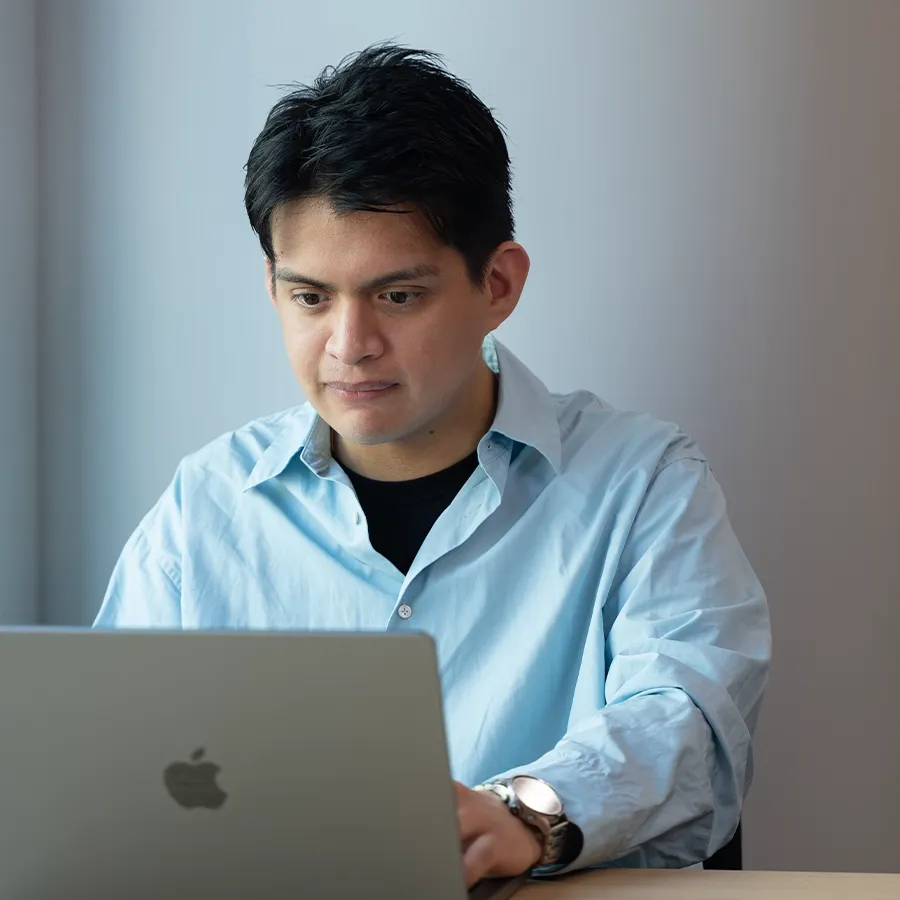 A man focused on his laptop screen, wearing a light blue button-down shirt in a bright room setting.