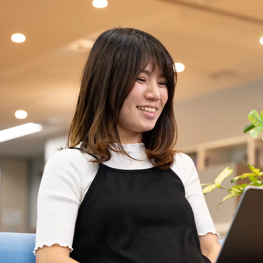 A smiling woman looking at a laptop in a modern, well-lit common area with indoor plants.